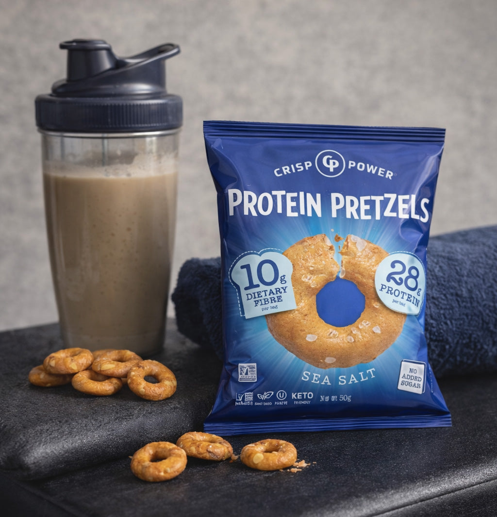 A bag of Crisp Power Protein Pretzels Sea Salt is displayed next to a shaker bottle with a protein shake and scattered pretzels on a gym bench.