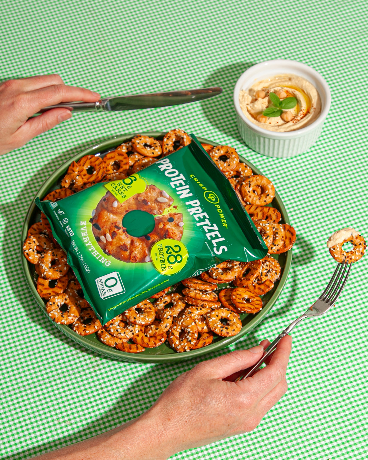 A green plate of protein pretzels topped with a bag of Crisp Power Everything Protein Pretzels, surrounded by hands holding a knife and fork on a green checkered tablecloth, with a small bowl of dip in the background.